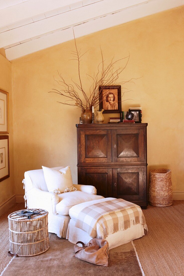 Sessel mit passendem Fussschemel neben Beistelltisch aus Rattan und Holzschrank an gelb getönter Wand im Dachgeschoss