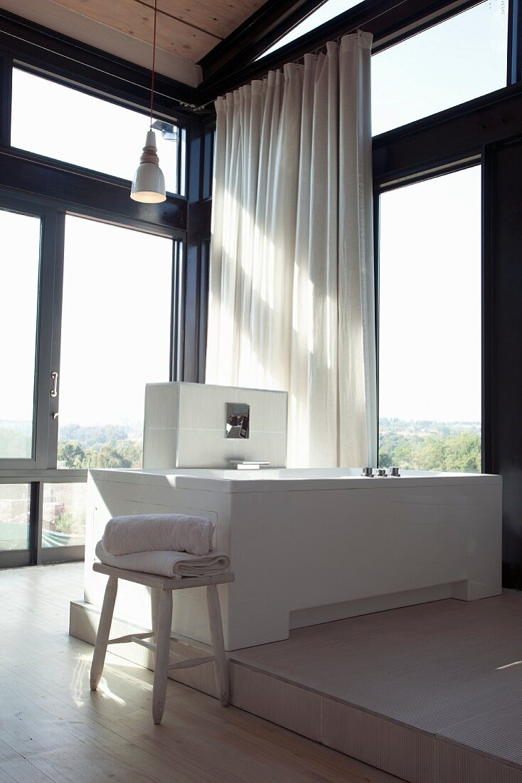 Badewanne auf Podest in verglaster Zimmerecke eines zeitgenössischen Wohnhauses und weisser Vorhang am Fenster