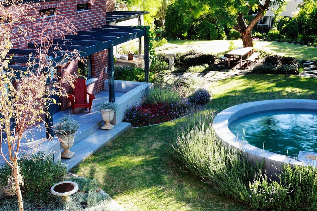 Rundes Wasserbecken im Garten vor Terrasse mit Pergola und Wohnhaus mit Ziegelfassade