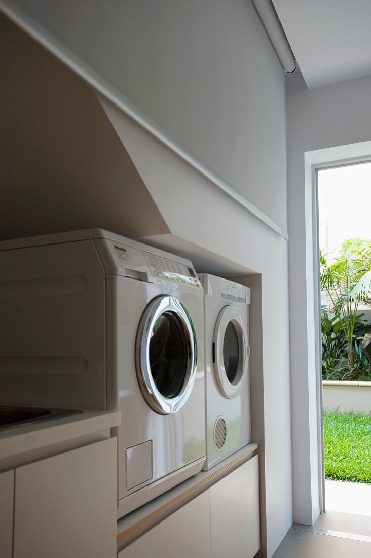 Wirtschaftsraum mit aufgeständerter Wasch- und Trockenmaschine neben offener Tür und Blick in sonnigen Garten