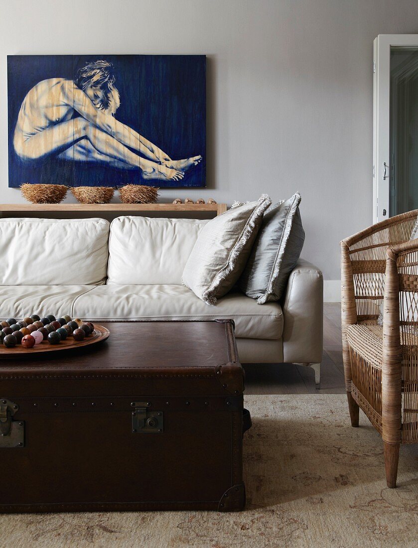 weiße Wohnzimmercouch mit darüberhängendem Aktgemälde; im Vordergrund eine antike Ledertruhe als Couchtisch und ein einfacher Rattan-Armstuhl