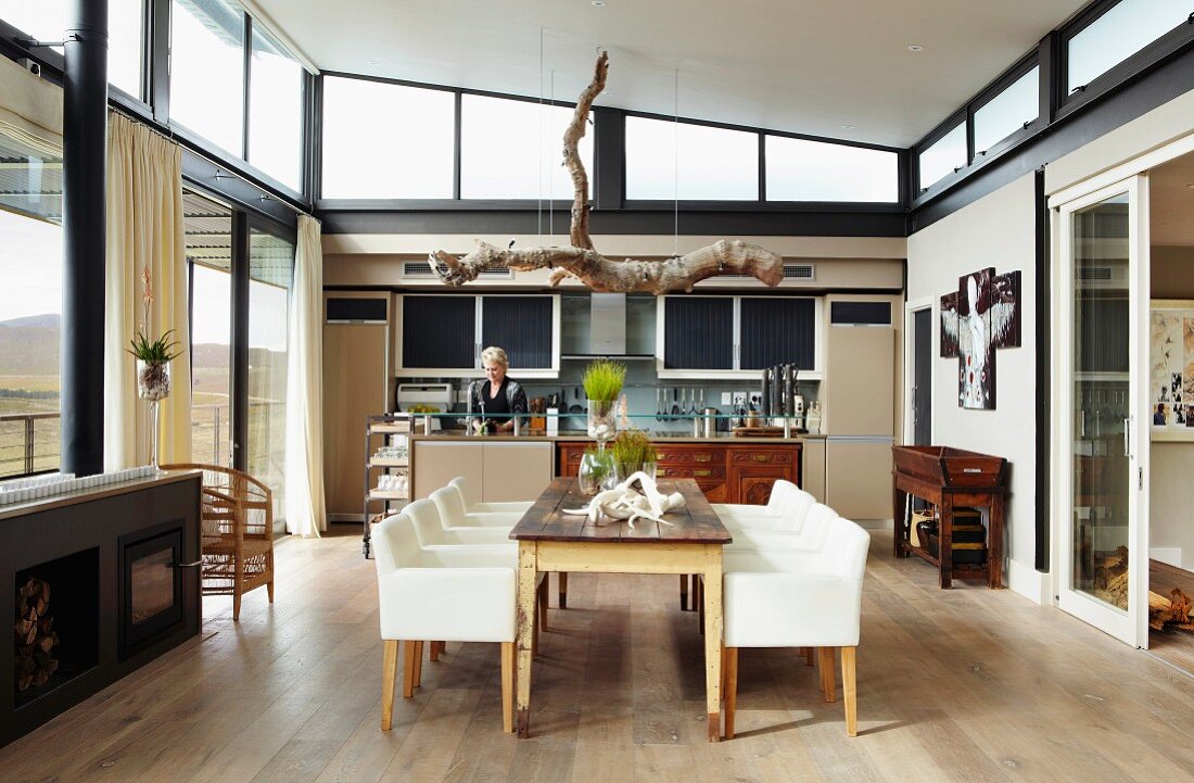 Modern kitchen counter with integrated antique elements; rustic dining table and elegant armchairs in foreground below pendant lamp made from large branch