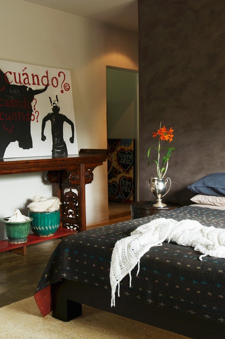 Schlafzimmer mit brauner Trennwand und antikem Wandtisch mit moderner Malerei; auf dem Nachttisch eine silberne Vase mit roten Blumen