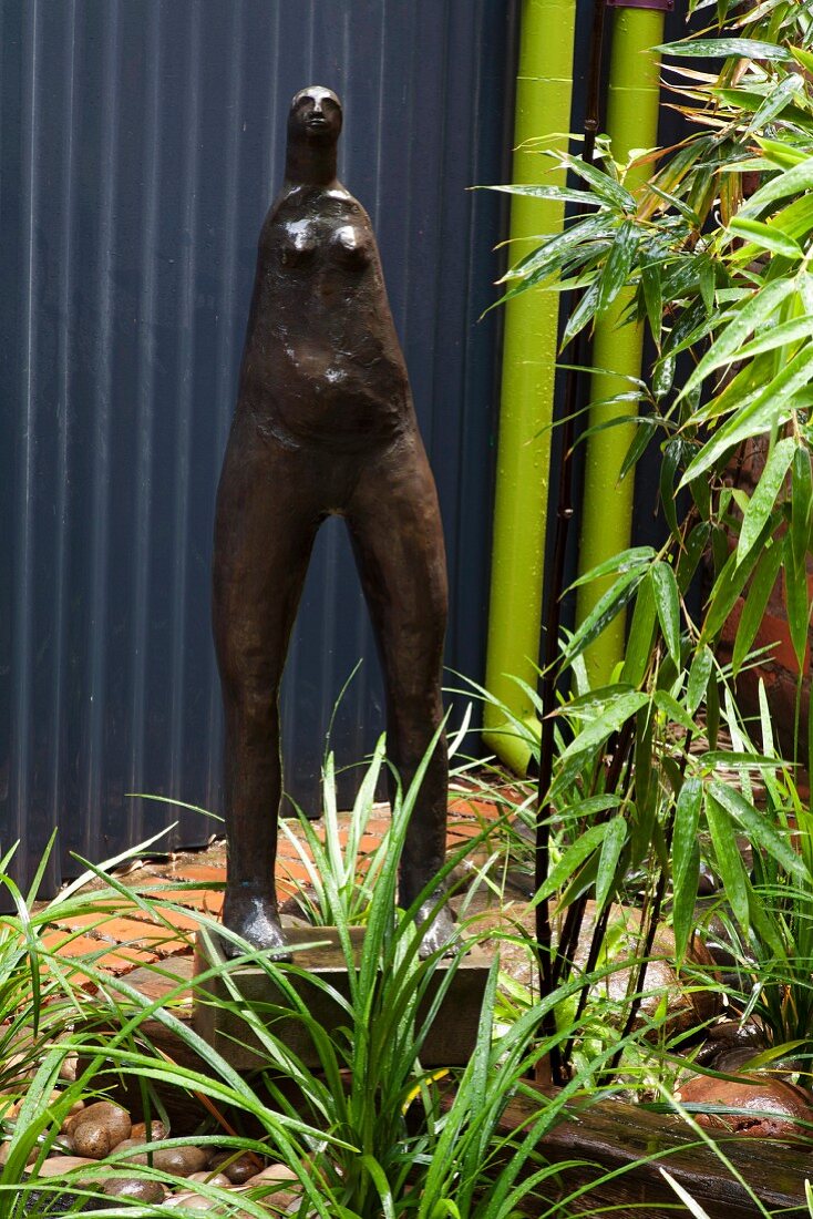Metall Frauenfigur auf kleinem Sockel im Garten vor Metall Hauswand und grünen Regenrohren