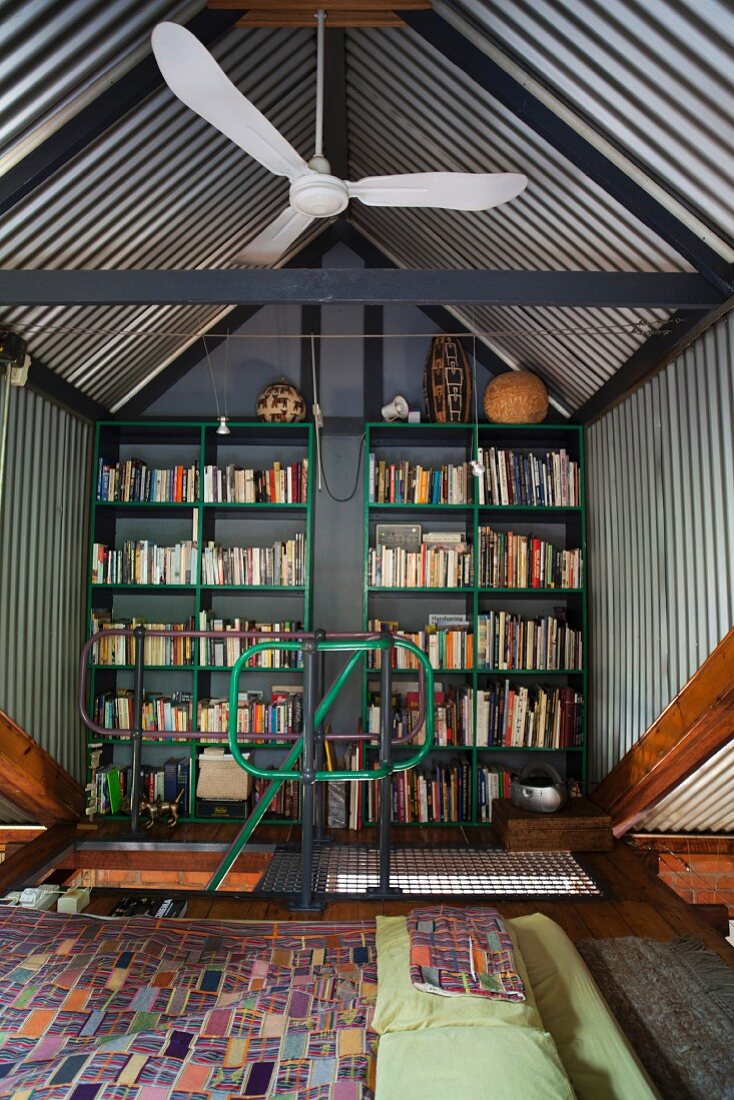 Sleeping area in front of bookcase on gable end wall below gable roof with corrugated metal cladding
