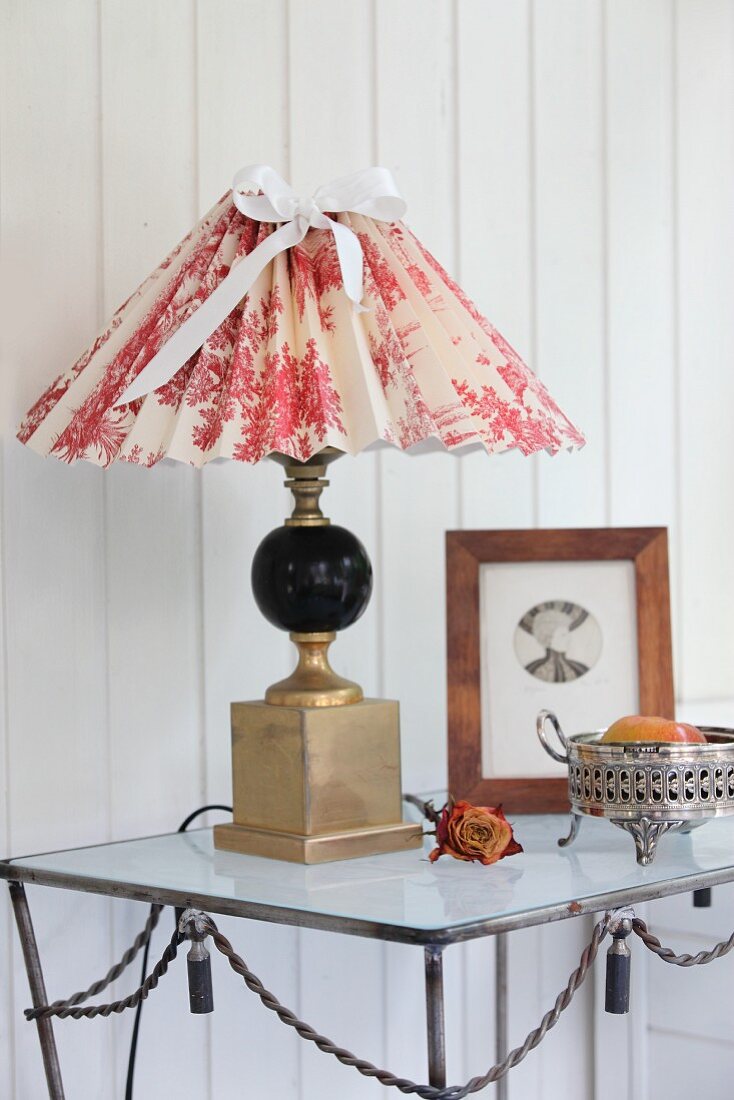 Table lamp with hand-crafted lampshade in red and white toile de jouy fabric on metal table against white, wood-clad wall