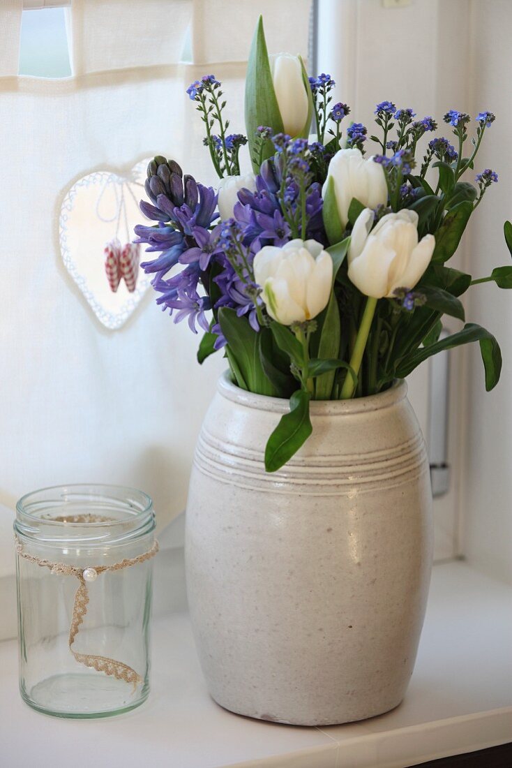 Frühlingsstrauss mit weissen Tulpen, blauen Hyazinthen und Vergissmeinnicht in schlichtem Keramikkrug vor Küchenvorhang mit Herzchenverzierung