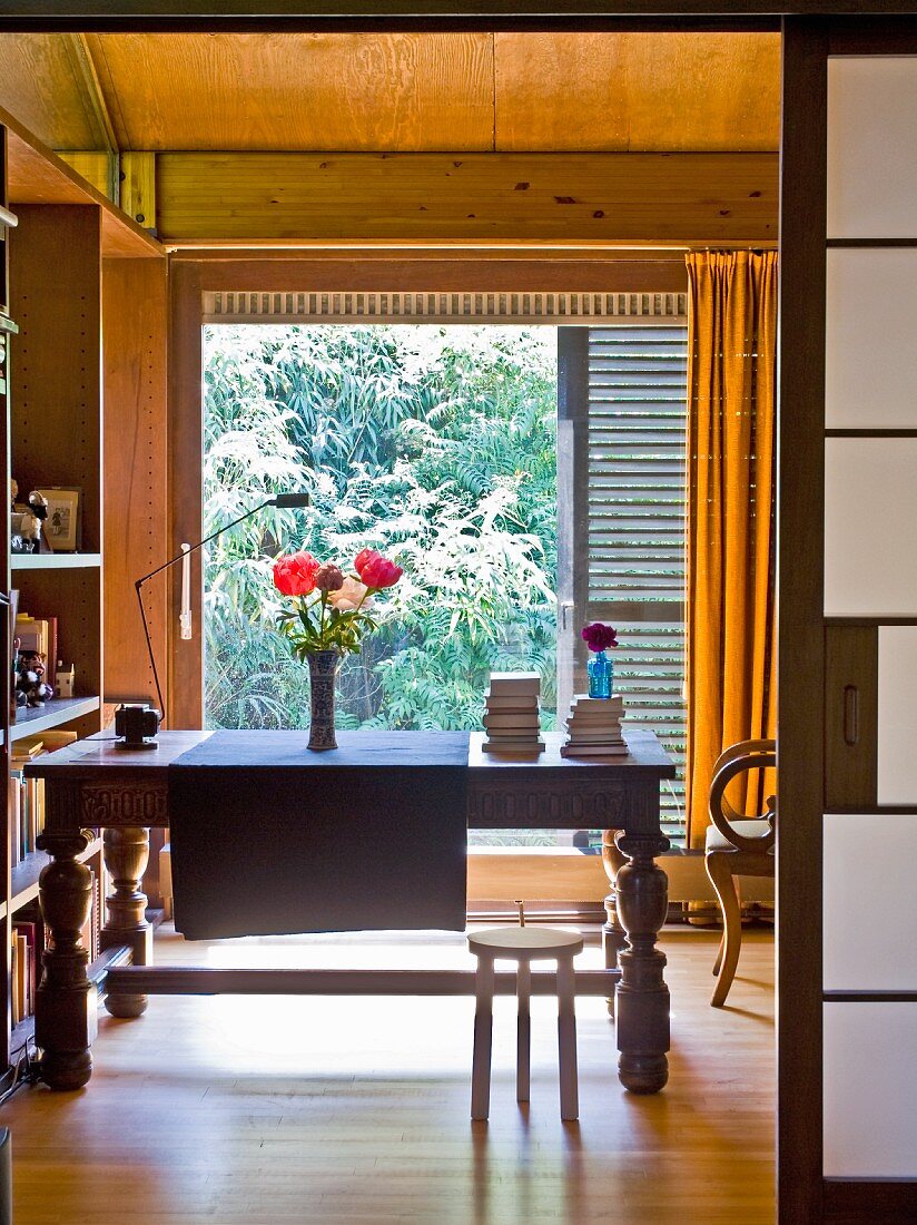 Blick durch offene Schiebetür auf antiken Holztisch vor raumhohem Fenster in holzverkleidetem Zimmer mit Gartenblick