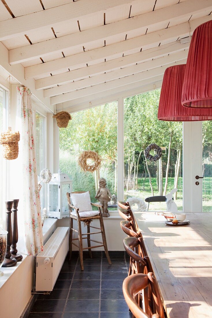 Sunny dining area in glazed extension with red pendant lamps hanging from white wooden ceiling