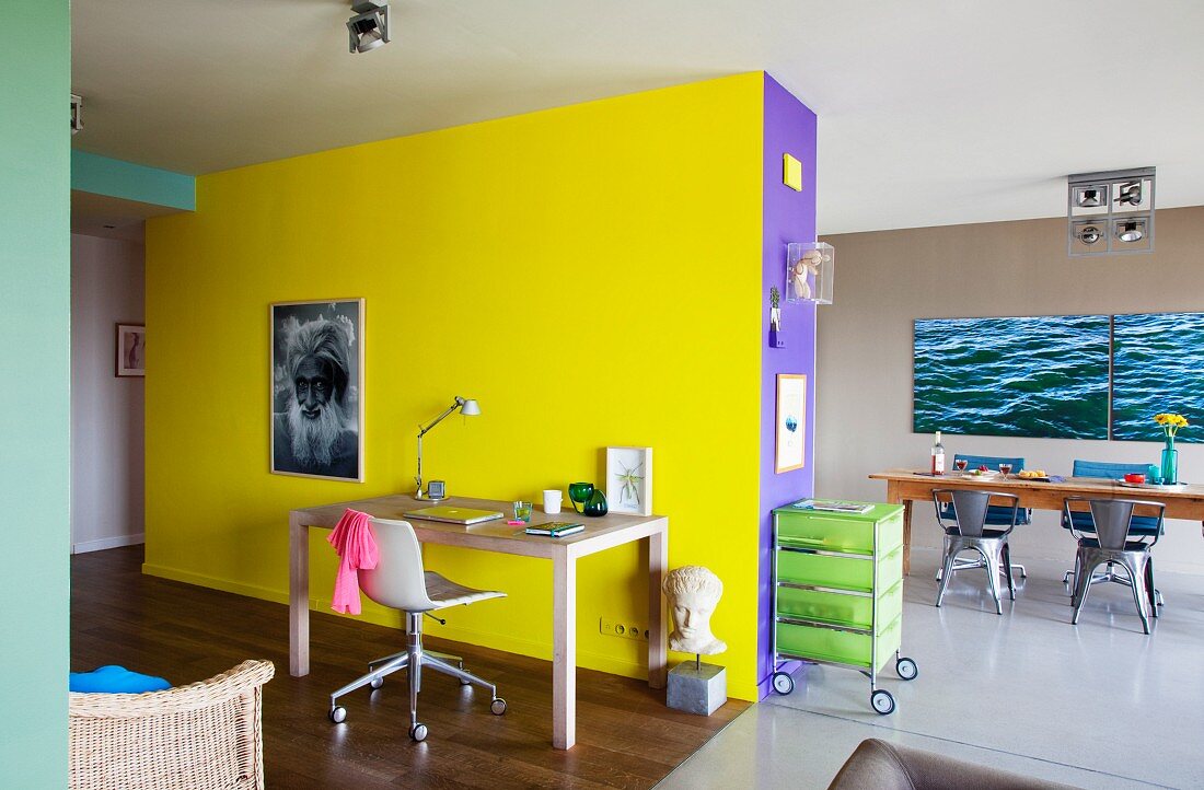 Desk and office chair against bright yellow wall in open-plan, colourful living area with dining table in background