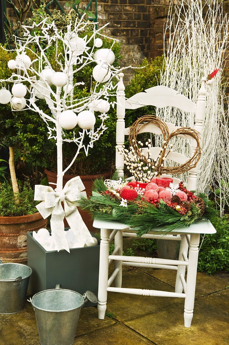 Weisslackiertes Bäumchen mit Kugel- und Sterndeko neben weihnachtlichem Gesteck auf Holzstuhl