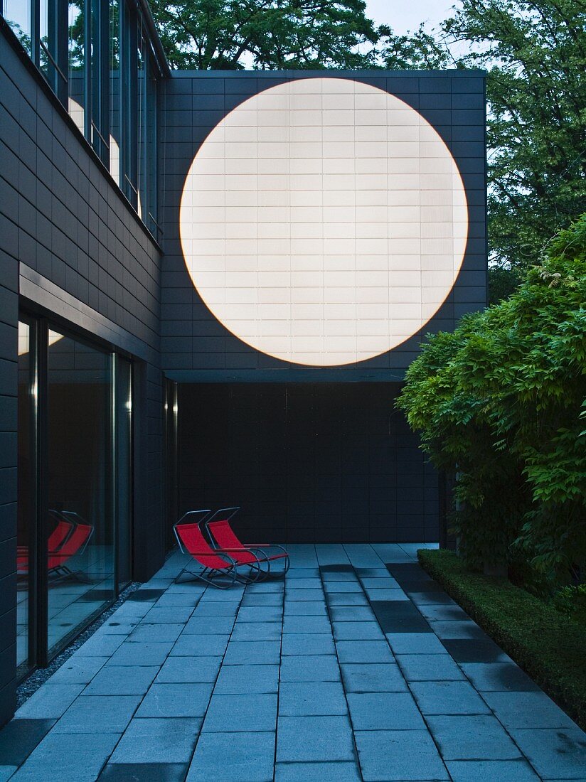 Anbau eines Wohnhauses mit grosser, weisser Kreisfläche auf dunkler Fassade in Abenddämmerung; auf Terrasse Liegestühle mit rotem Bezug