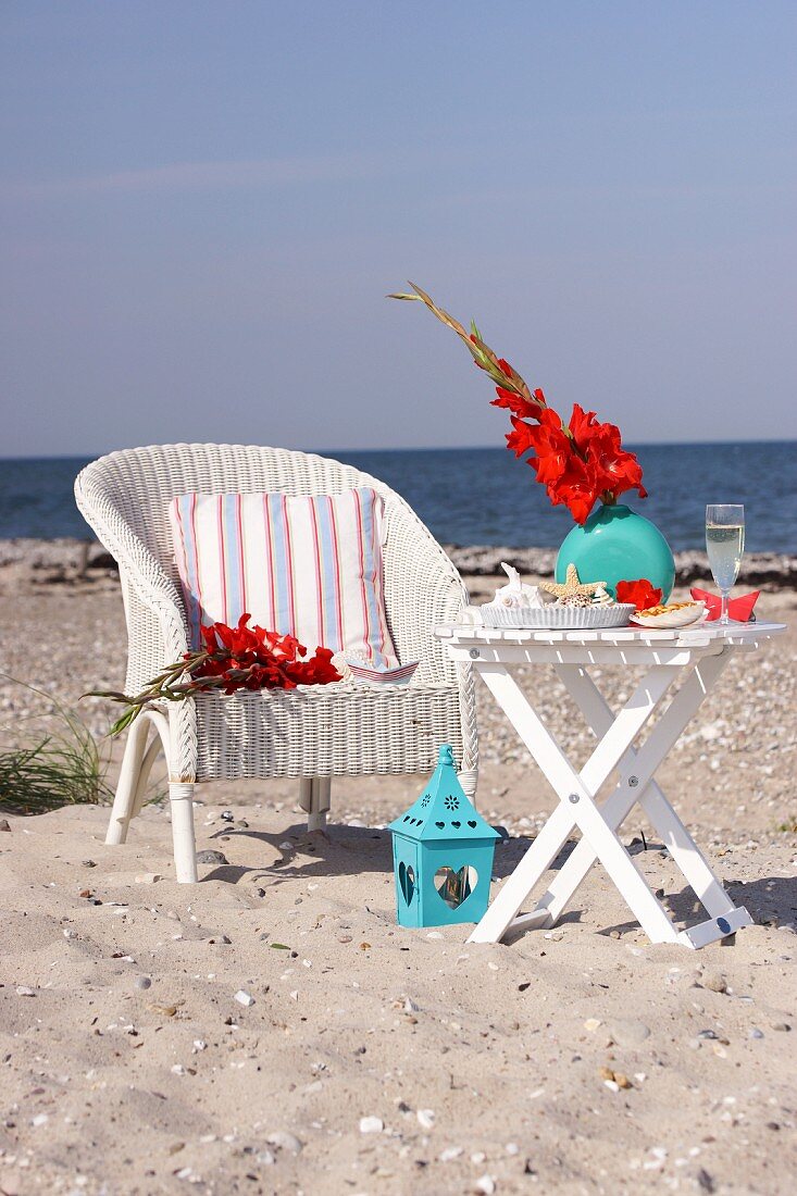 Vase of gladioli, maritime ornaments and glass of champagne on folding table next to wicker chair on sandy beach