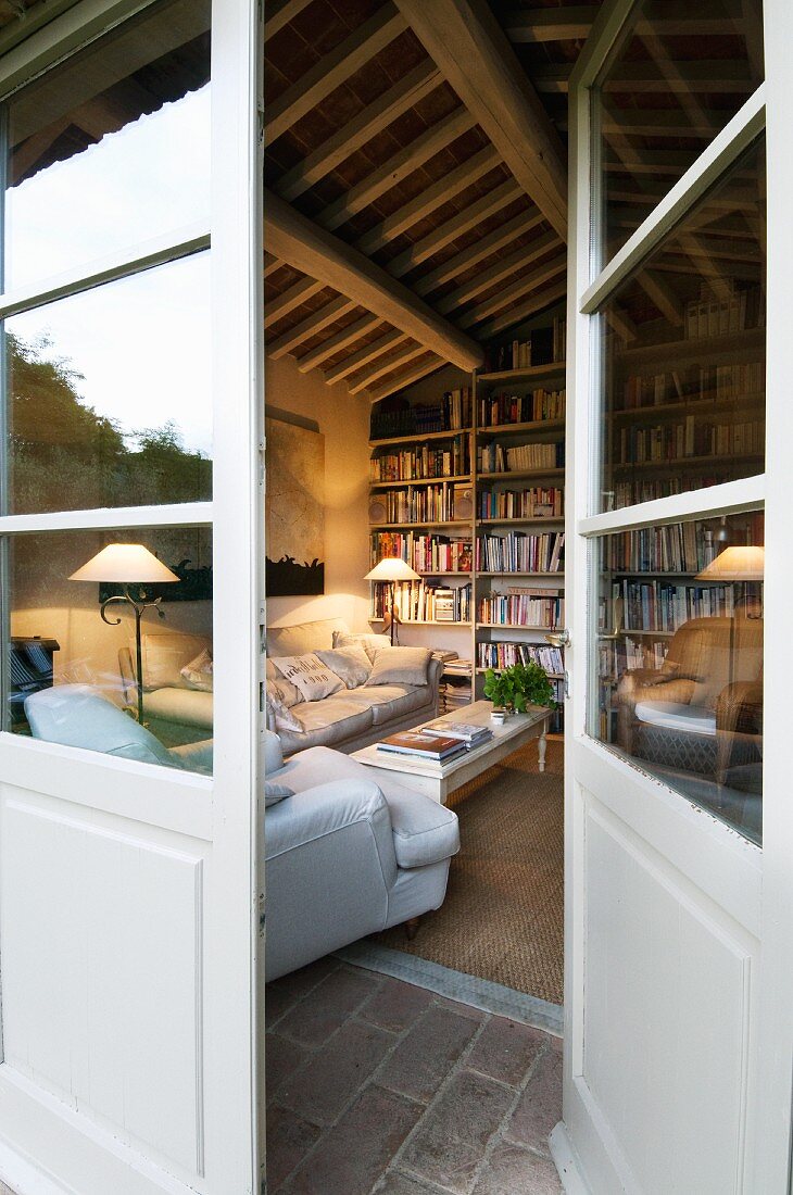 View through open French doors into comfortable living room with large bookcase below rustic wooden ceiling