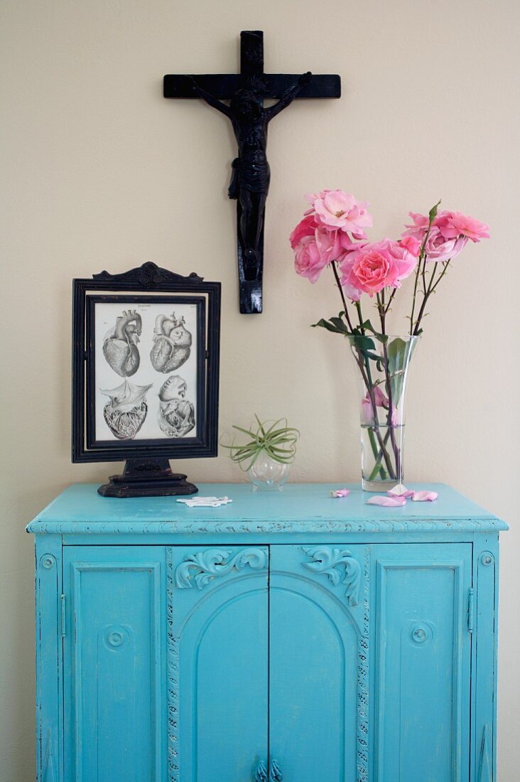 Pink roses and framed anatomical drawing on blue wall cabinet below crucifix