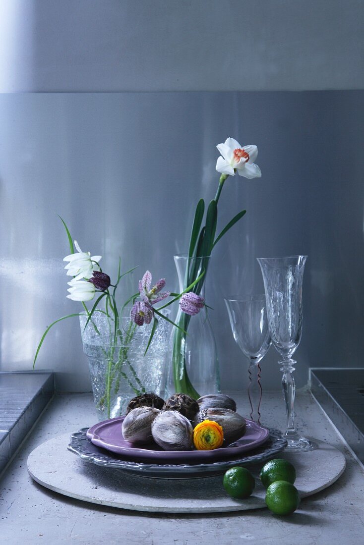 Stillleben mit Deko-Ostereiern auf Schalen, Gläsern und Frühlingsblumen auf grauem Betongrund vor grau getönter Wand