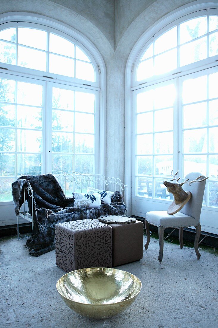 Shiny brass bowl on floor in front of cubic pouffes and vintage upholstered chairs in corner of room with arched French windows