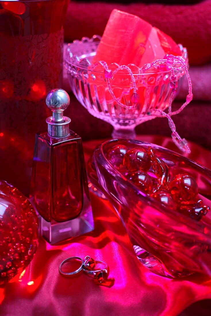 Still-life arrangement in red - perfume bottle next to various glass dishes on red satin cloth