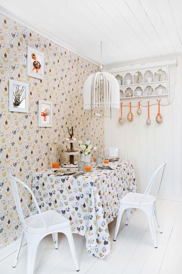 Dining area set for Easter in kitchen decorated with hen motif on wallpaper and tablecloth and white, vintage chairs