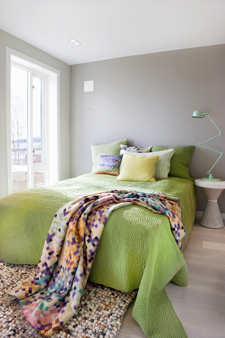 Leuchtenklassiker auf Beistelltisch neben einem Bett mit lindgrünem Überwurf und grafisch gemustertem Plaid