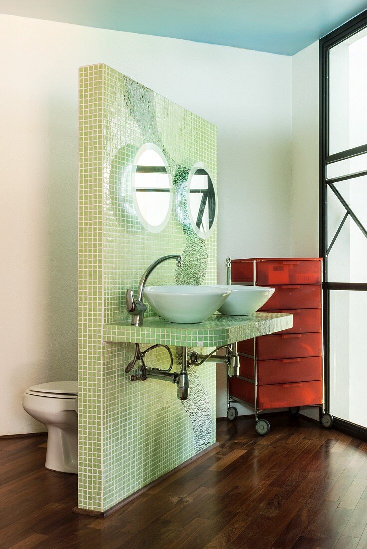 Zum Fenster gewandter Doppelwaschtisch und Toilette an der Rückseite einer freistehenden Wand mit lindgrünen Mosaikfliesen