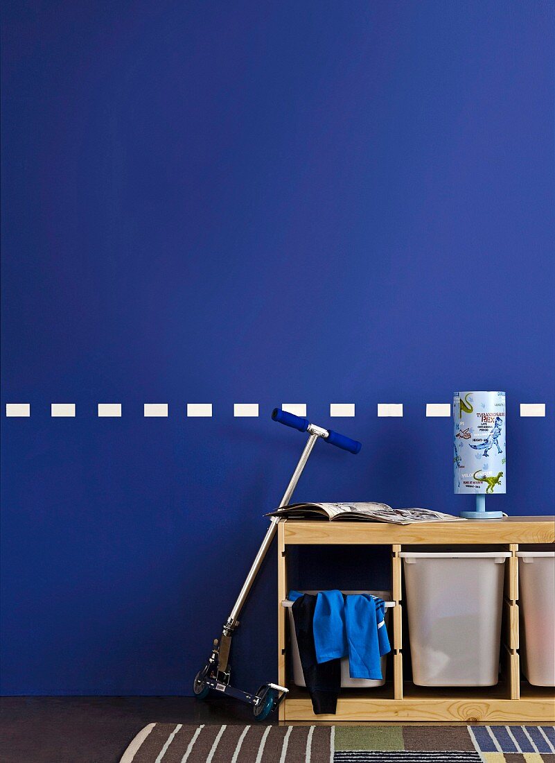 Praktisches Aufbewahrungsmöbel im Kinderzimmer mit Dino-Lampe und angelehntem Roller vor blauer Wand