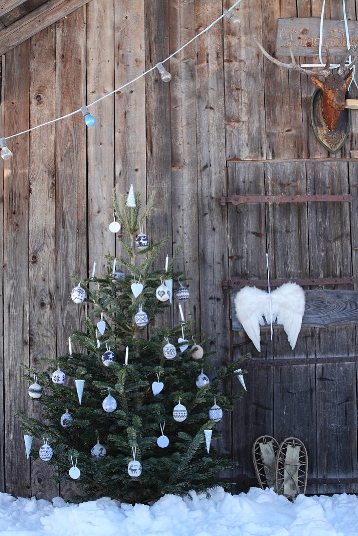 Geschmückter Weihnachtsbaum mit selbst gestrickten Kugeln und Engelsflügel an rustikaler Holzhütte im Schnee