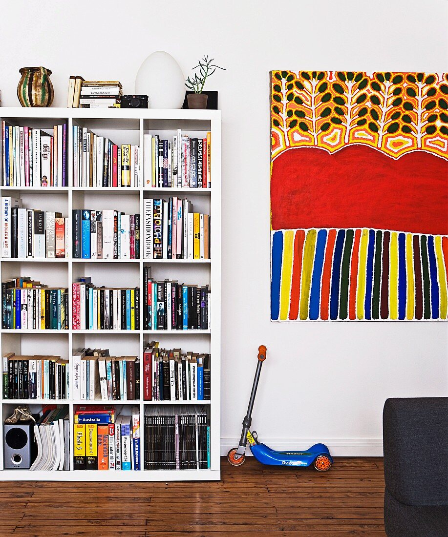 White bookcase with square divisions next to child's scooter and modern, colourful artwork on wall (Aboriginal art)