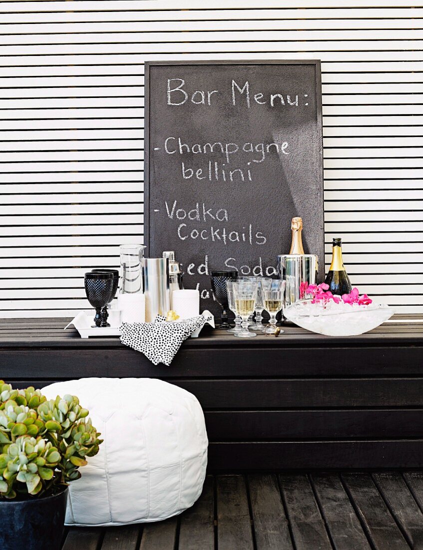 Improvisierte Bar mit Gläsern und Flaschen auf dunkler Holzbank, Schiefertafel mit Beschriftung an weisser Lamellenwand lehnend