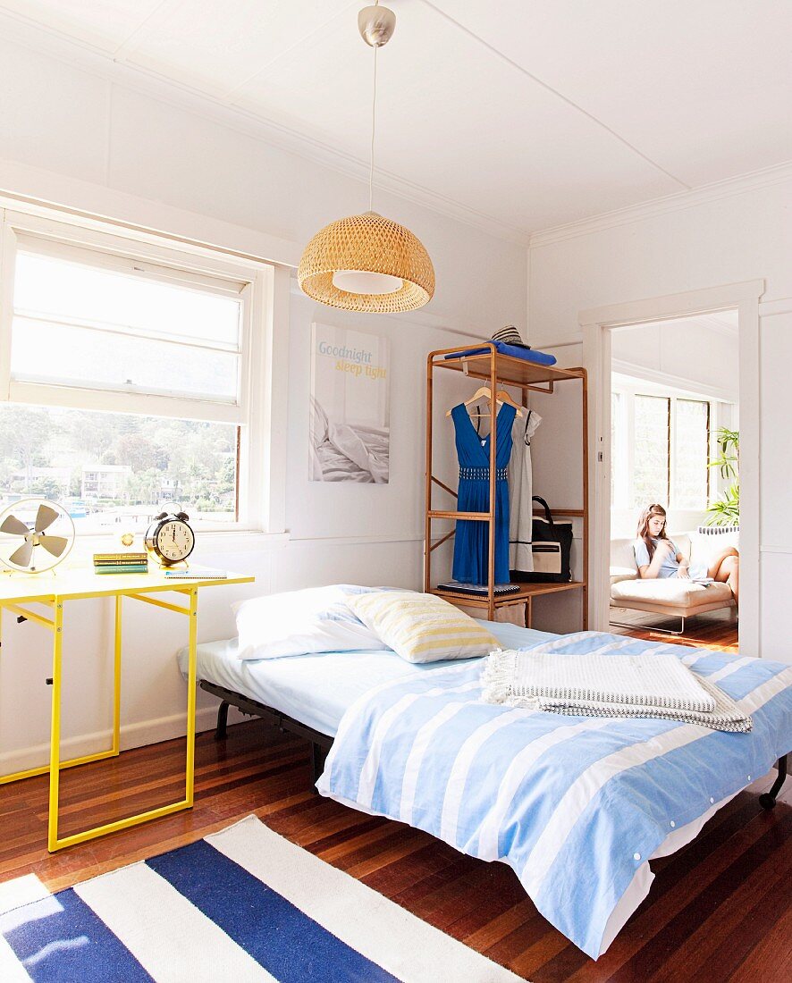 Bett mit schlichtem Metallgestell und gestreifte Bettwäsche neben Schreibtisch mit gelbem Metallgestell am Fenster, im Hintergrund offene Tür und Blick auf Mädchen im Sessel