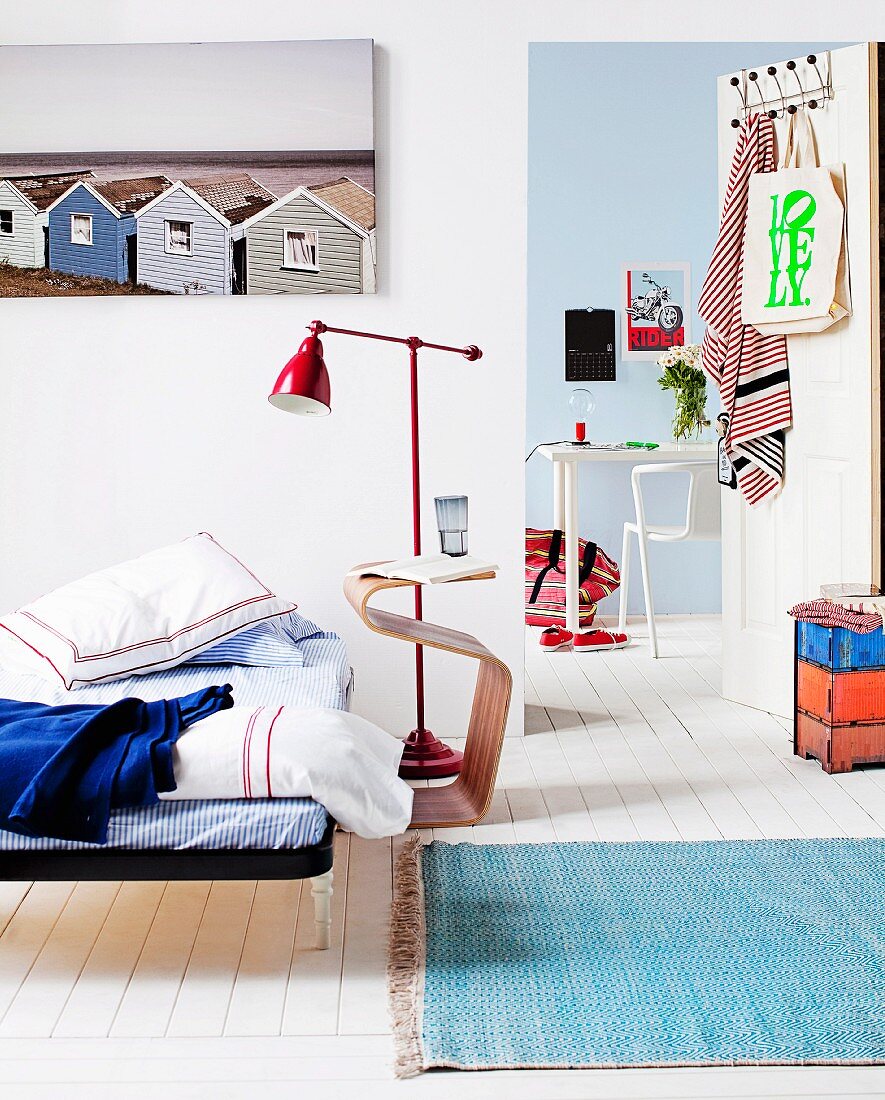 Schlichtes Bett mit Kissen neben modernem Beistelltisch aus Holz vor Retro Stehleuchte aus rotem Metall, Blick durch offene Tür auf modernem Arbeitstisch in ländlichem Ambiente