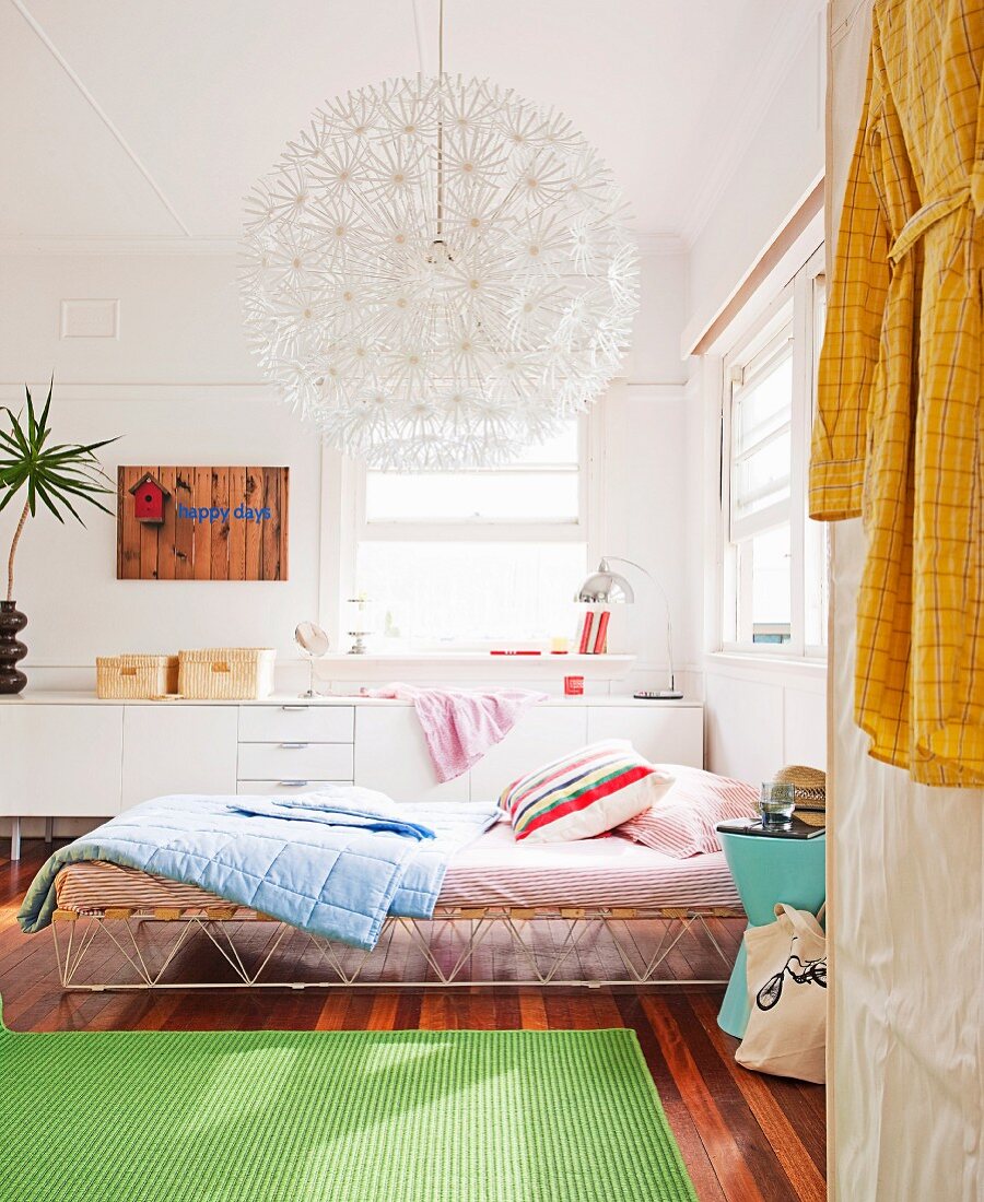 Grüner Teppich auf Holzboden vor Bett mit Retro Metallgestell und Kissen unter kugelförmiger Hängeleuchte aus einzelnen Blumenelementen, im Hintergrund weisses Sideboard