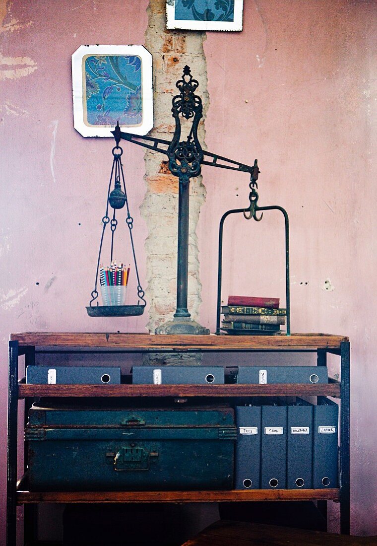 Antique scales on simple, half-height shelving with metal trunk and blue box files against pink wall with damaged plaster