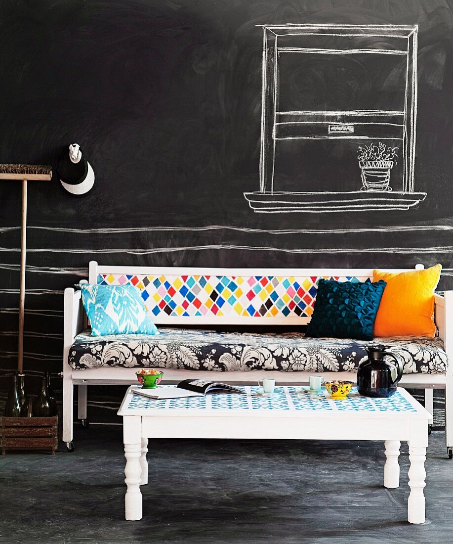 Weiß lackierte Holzbank und Tisch mit buntem Stoffmix; Wand und Boden mit Tafelfarbe gestrichen