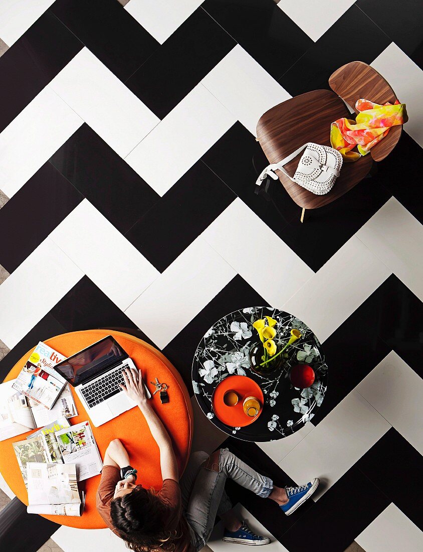 Frau vor orangem Polsterhocker mit Laptop und Arbeitsunterlagen in Wohnraum mit dekorativen Fliesenboden aus grossformatigen schwarz-weißem Zickzackmuster