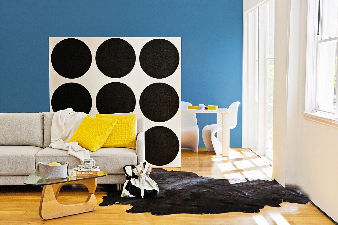 Seating area with sofa, animal-skin rug & coffee table separated by partition shelving with back wallpapered in black and white pattern of circles