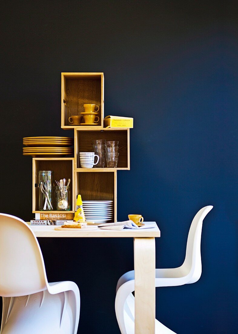 White, classic shell chairs at wooden table with crockery in shelving modules against blue wall