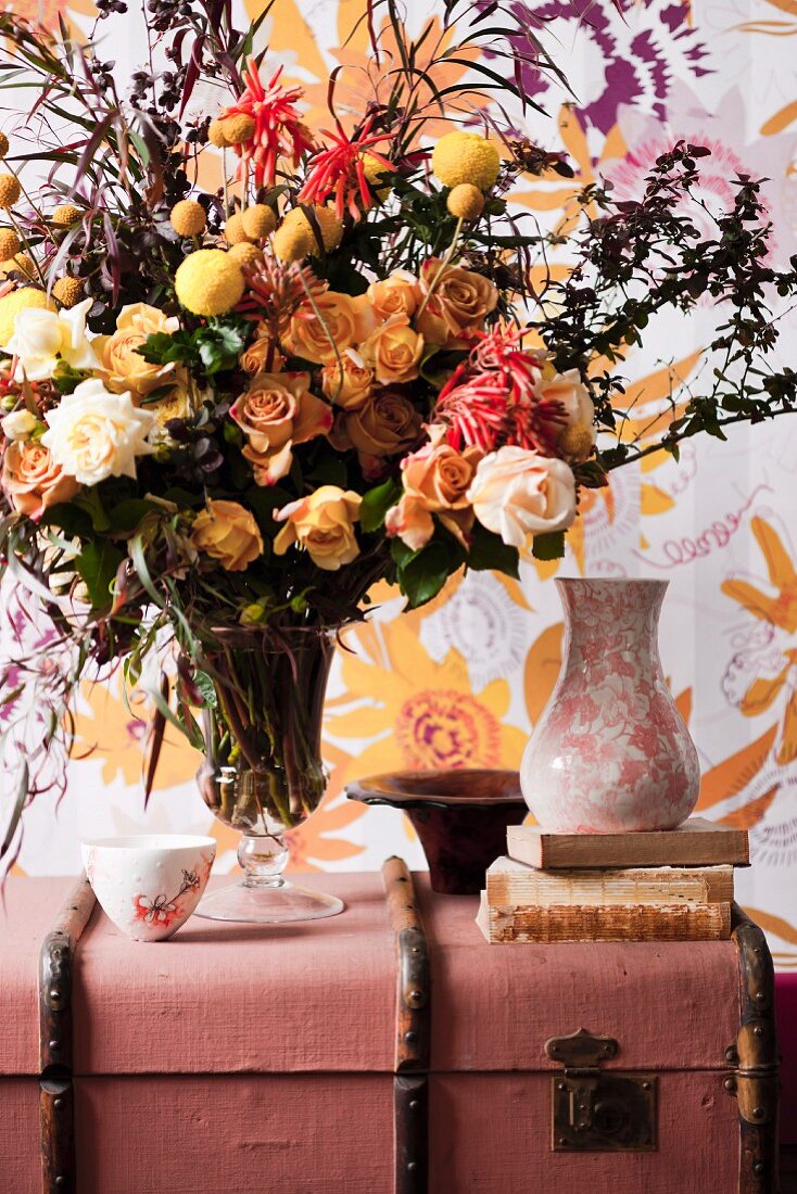 Üppiger Blumenstrauss in Glasvase und Keramikvase auf antikem, rosa Koffer, im Hintergrund geblümter Wandbehang