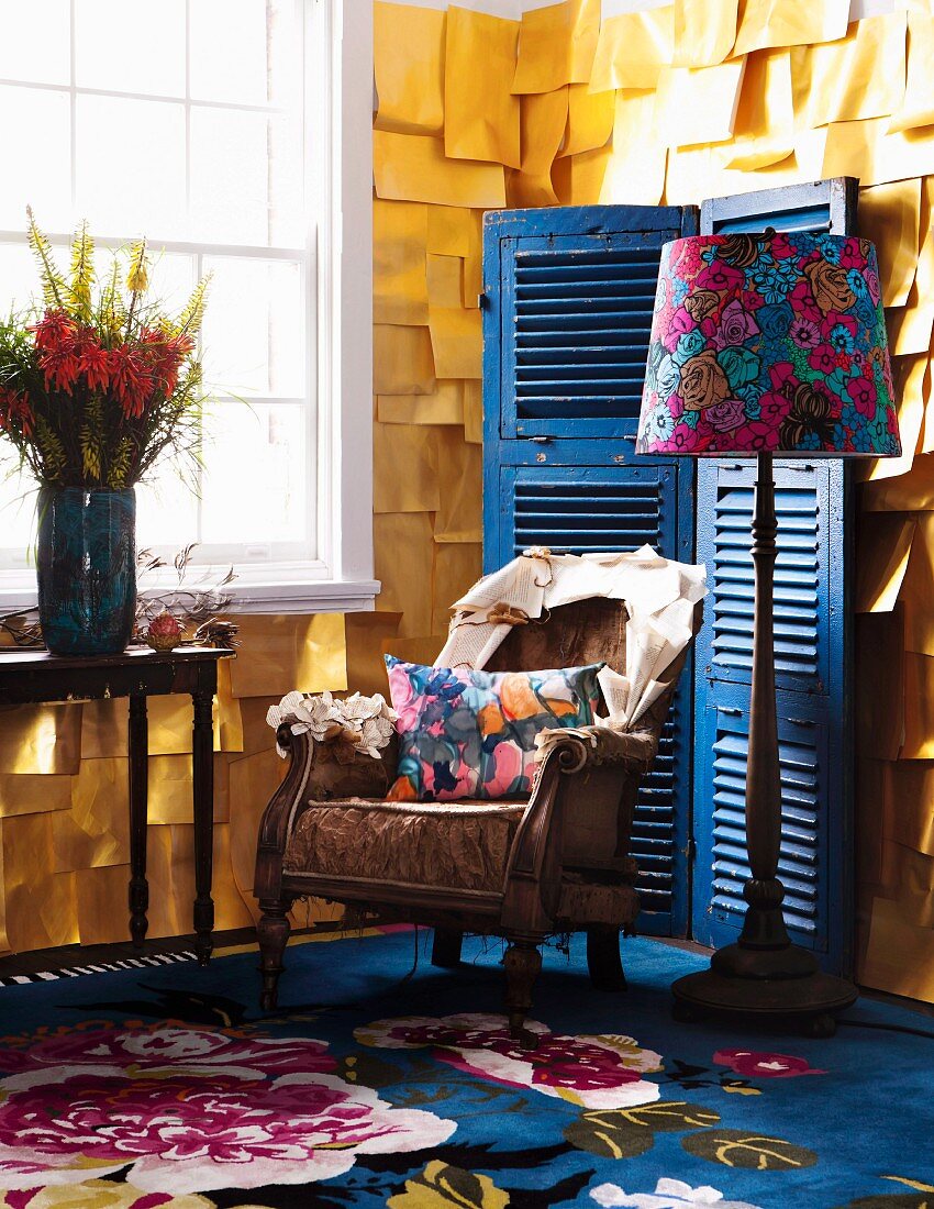 Vintage Sessel mit Kissen, Stehleuchte mit geblümten Schirm und Blumenstrauss auf Konsolentisch vor Sprossenfenster, an Wand gelbe Papier Blätter schuppenartig befestigt