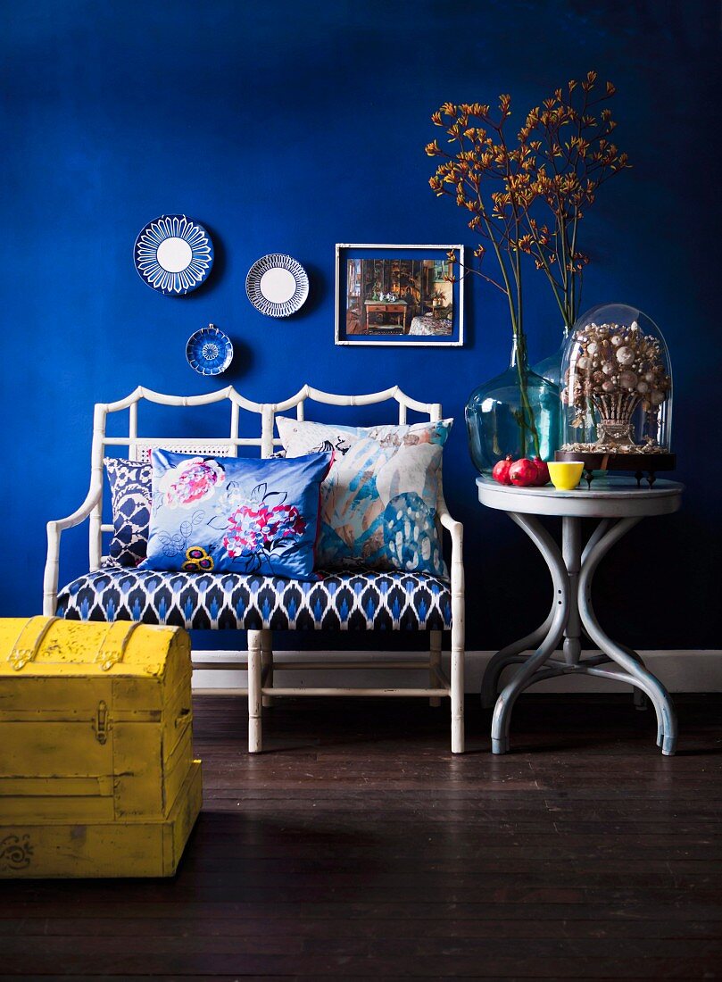 weiße Rattan Sitzbank mit gemustertem Polsterbezug und Kissen, daneben runder Beistelltisch mit Blumenzweigen in Glasvase vor blau getönter Wand, seitlich teilweise sichtbare gelbe Holztruhe