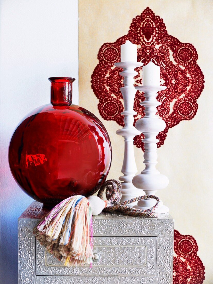 Dekoratives Arrangement mit roter Glasvase und weißen kunsthandwerklichen Kerzenständern vor Ornamenttapete