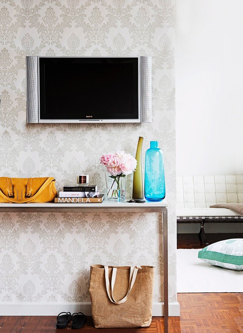 TV mounted on white brocade wallpaper above various vases and mustard yellow handbag on simple, narrow console table