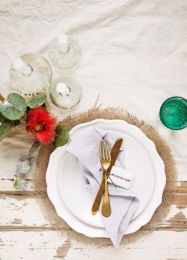 Weißes Teller mit goldfarbenem Besteck und Stoffserviette auf Shabby-Chic Holztisch dekoriert mit weißen Kerzen und roter Dekoblume