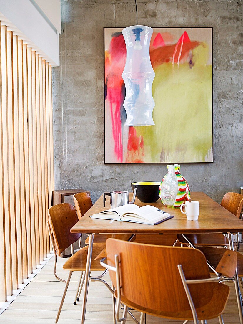 Essplatz mit Holzstühlen und passendem Tisch im Fiftiesstil gegenüber Wand mit modernem Bild, seitlich vertikale Holzlamellen vor Fenster