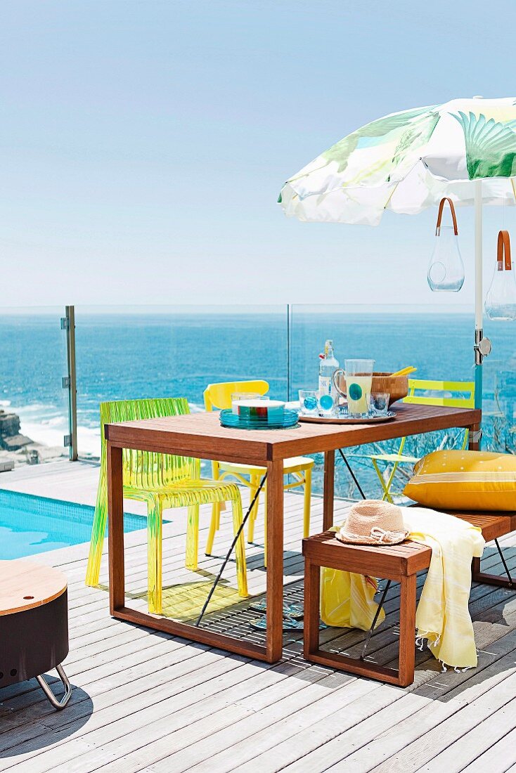 Holztisch mit passender Bank und gelben Stühle auf sonnenbeschienener Holzterrasse am Pool mit Blick über Glasbrüstung auf das Meer