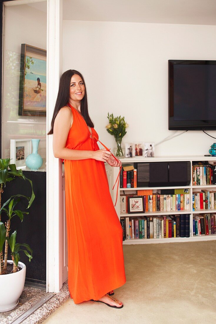 Woman wearing long orange dress leading on terrace door frame in front of half-height white bookcases below wall-mounted TV