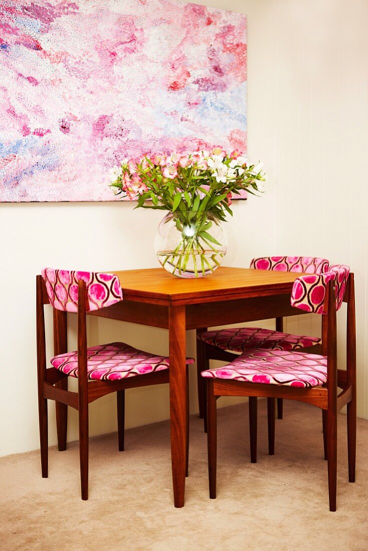 Holzstühle mit gemustertem Stoffbezug auf Sitzpolster und Rückenlehne um Tisch mit Blumenstrauss, an Wand grossformatiges Bild