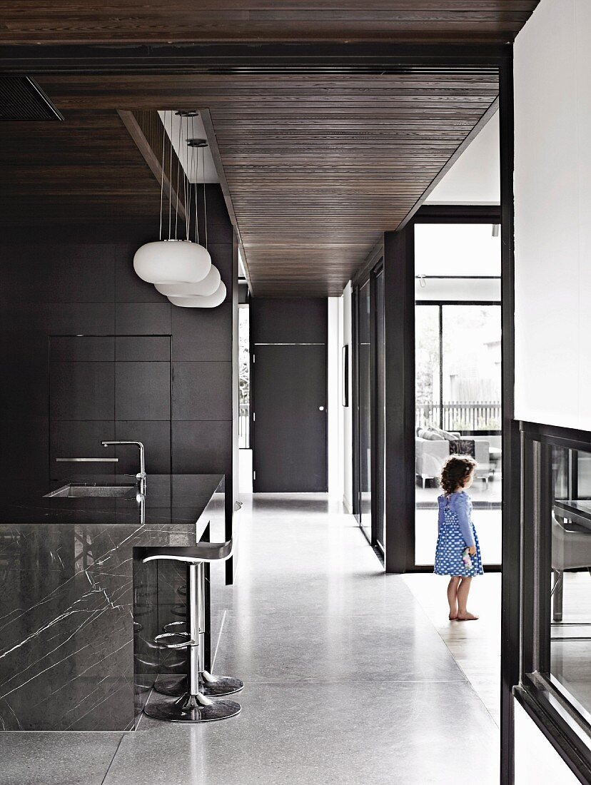 Gangflucht in minimalistischem Wohnhaus mit Wandelementen und Fensterrahmen in dunklem Grau, seitlich natursteinverkleidete Kücheninsel mit Barhockern
