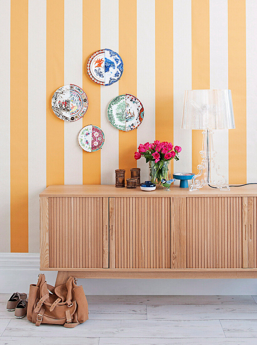 Designer plastic table lamp on light wooden sideboard, wall plate on yellow and white stripe wallpaper