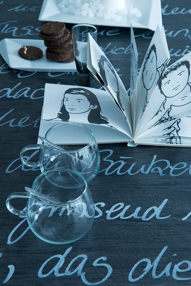 Glass cups and book of black and white drawings on blackboard tabletop with white lettering